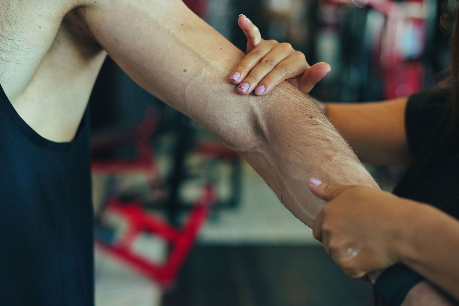 Person's arm being held by another's hands. One is wearing a black tank top, in a gym setting.