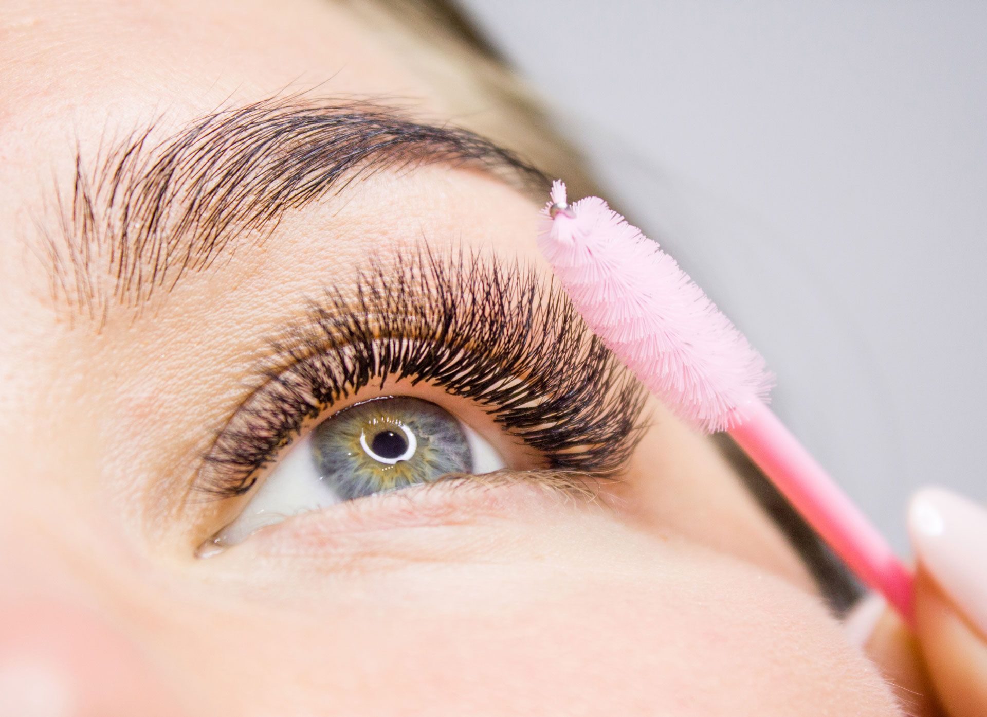 Close-up of eye with long, dark lashes being brushed with a pink lash wand. Eye is blue-green.