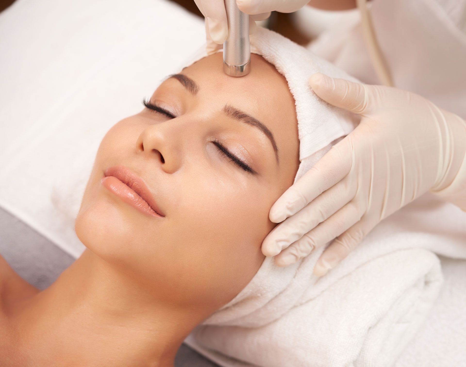 Woman receiving facial treatment, hands holding tool to face, eyes closed, spa setting.