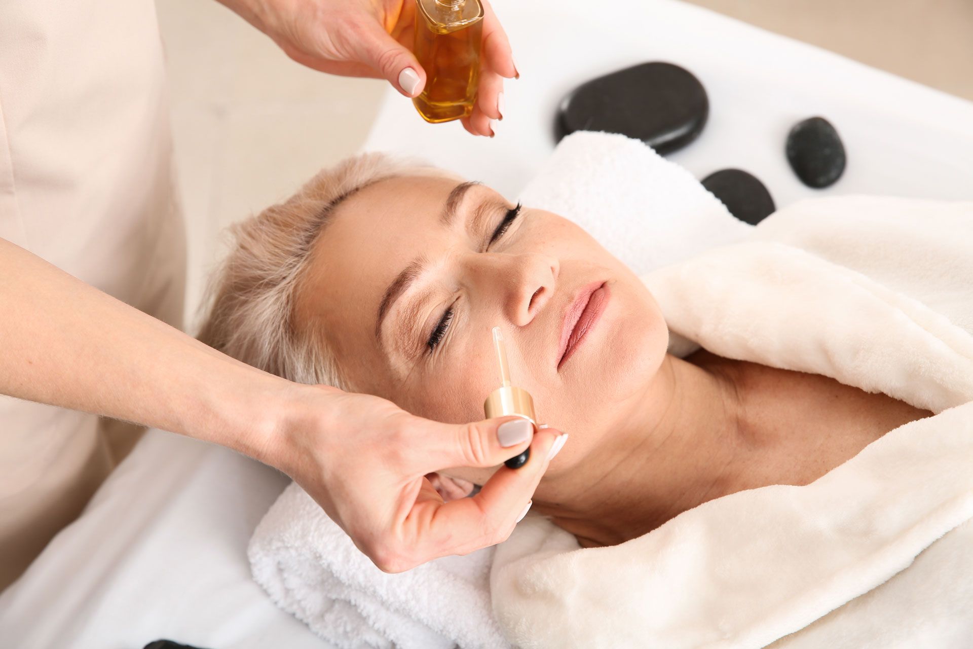 Woman receiving facial treatment; oil being applied with wand in spa setting.