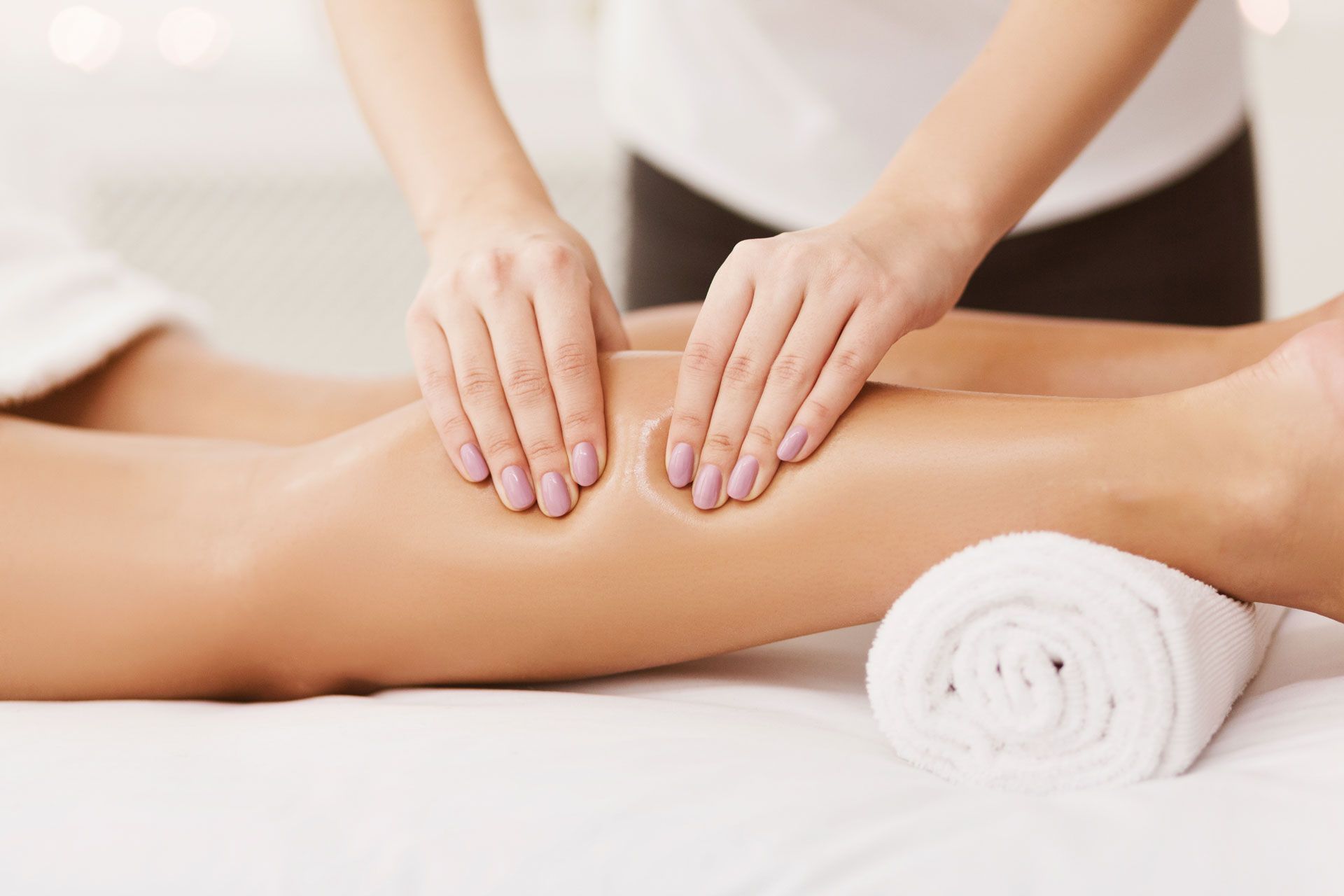 Hands massaging a leg on a white surface with a rolled towel, possibly in a spa setting.