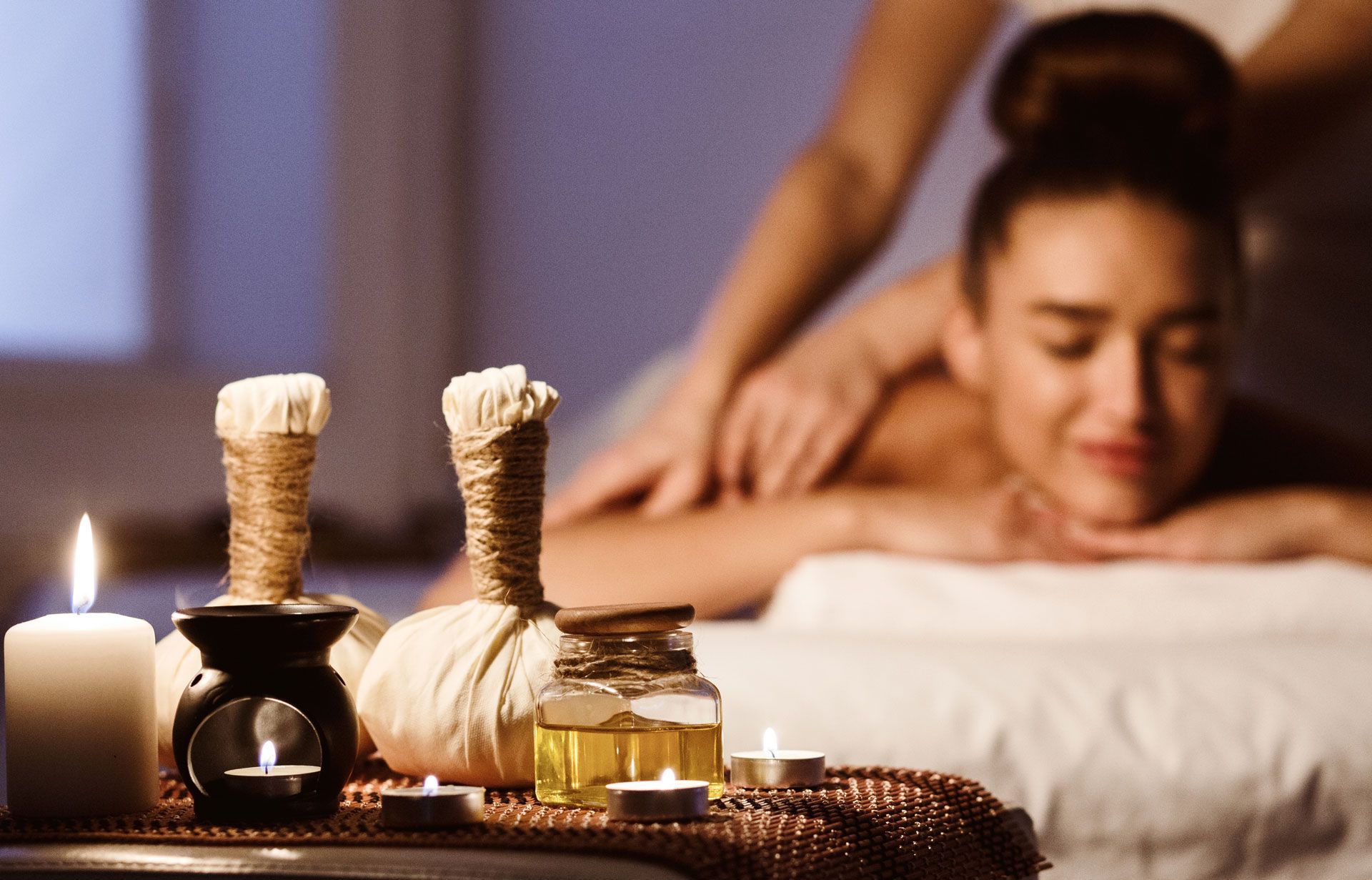 Spa scene: Woman receiving massage, candles, oil, and herbal compress on a table.