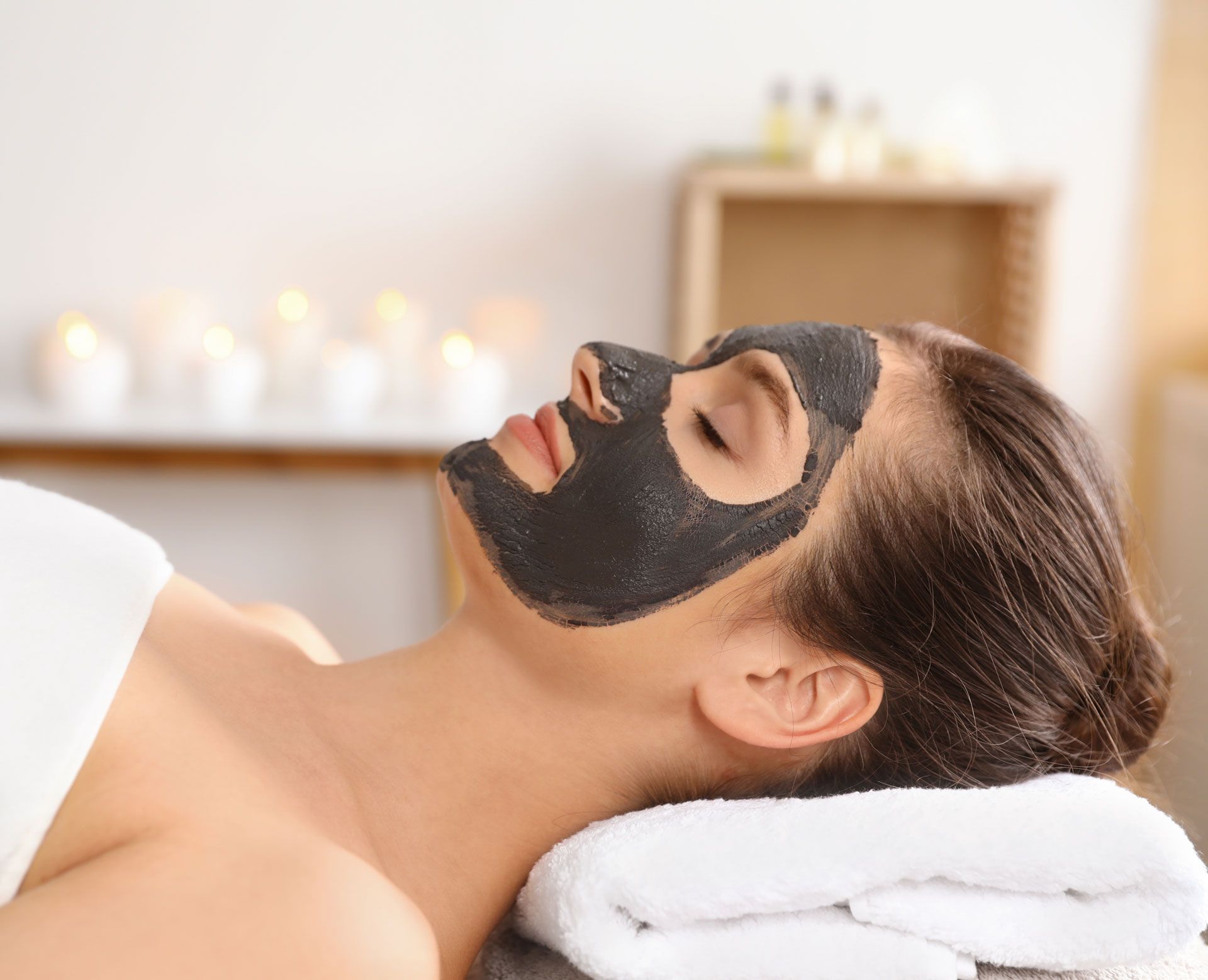 Woman with a black facial mask relaxes on a massage table, spa setting.