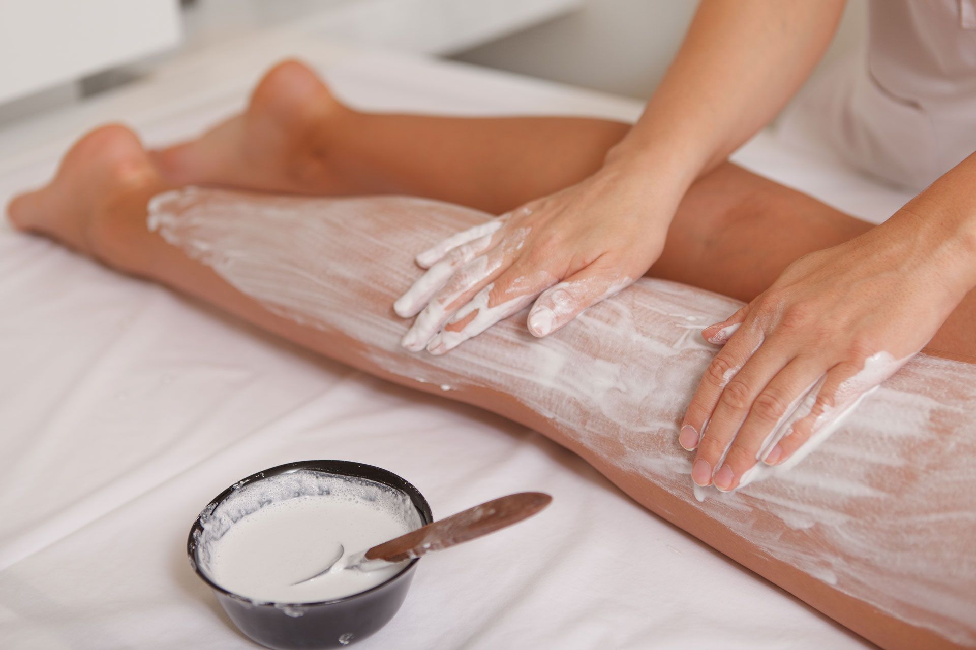 Person applying white cream to a leg on a white surface, with a bowl of cream nearby.