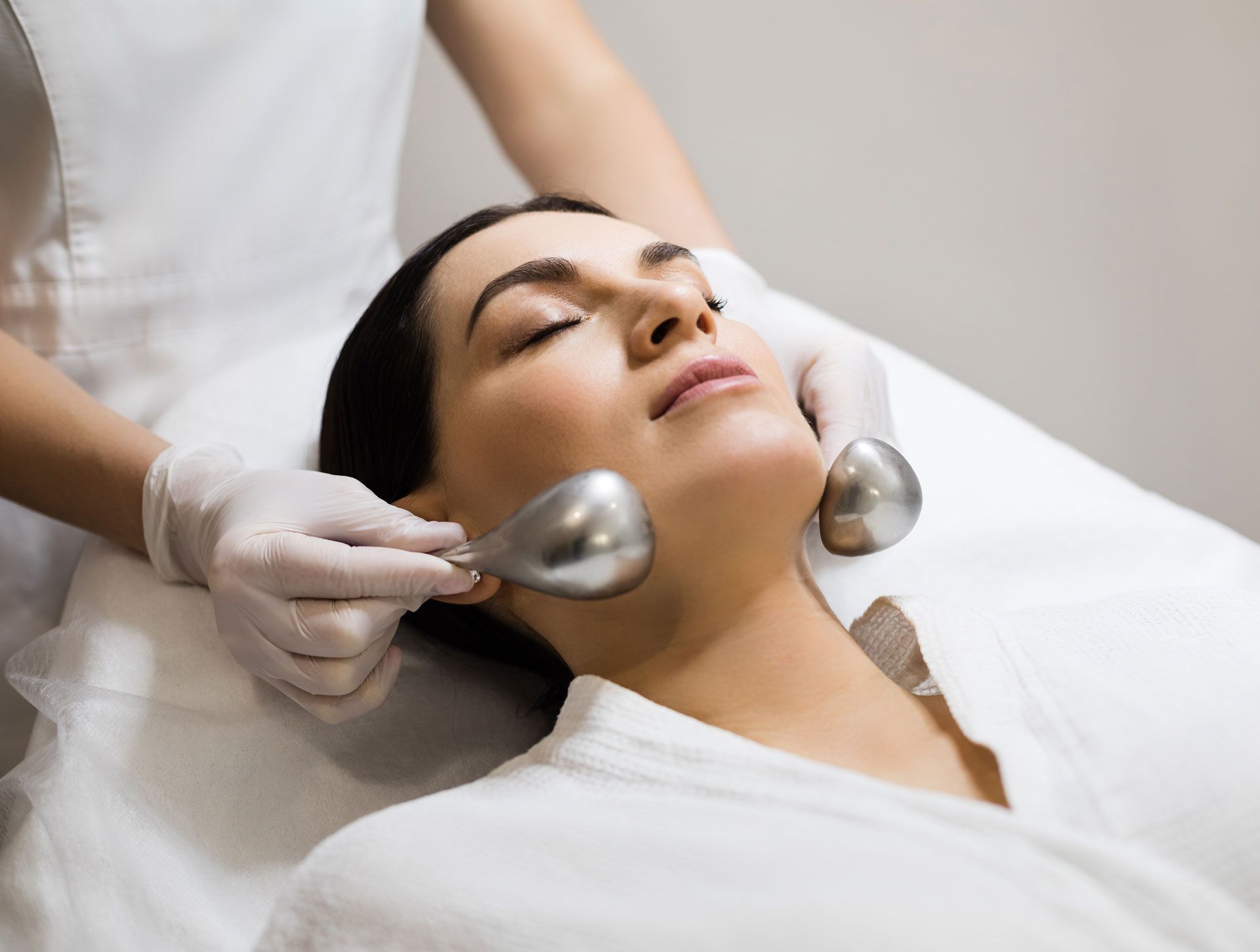 Woman receiving facial treatment with metal ice globes. She has eyes closed, laying on a white surface.