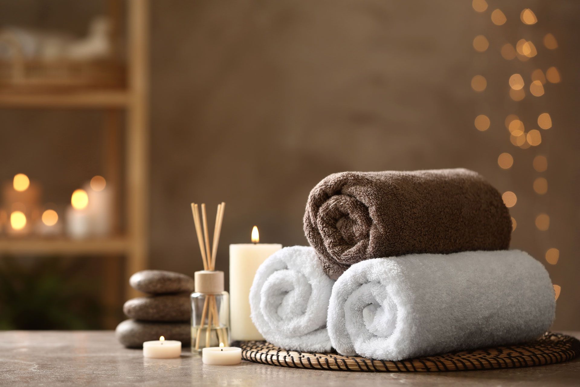 Rolled towels, candles, aroma diffuser, and massage stones arranged on a table for spa ambiance.
