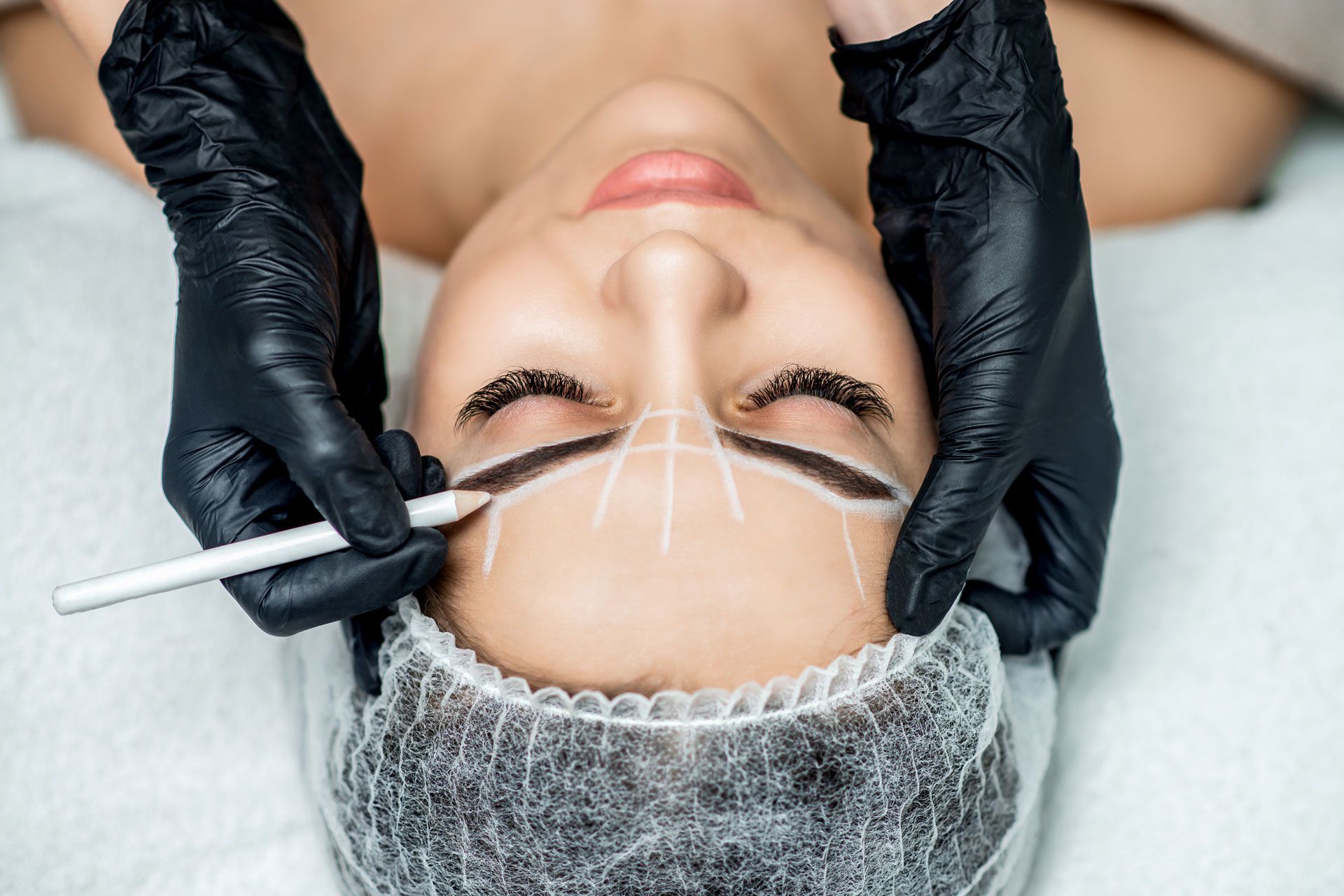 Person having eyebrows mapped with a pencil before a cosmetic procedure, wearing a hair net, with gloved hands.