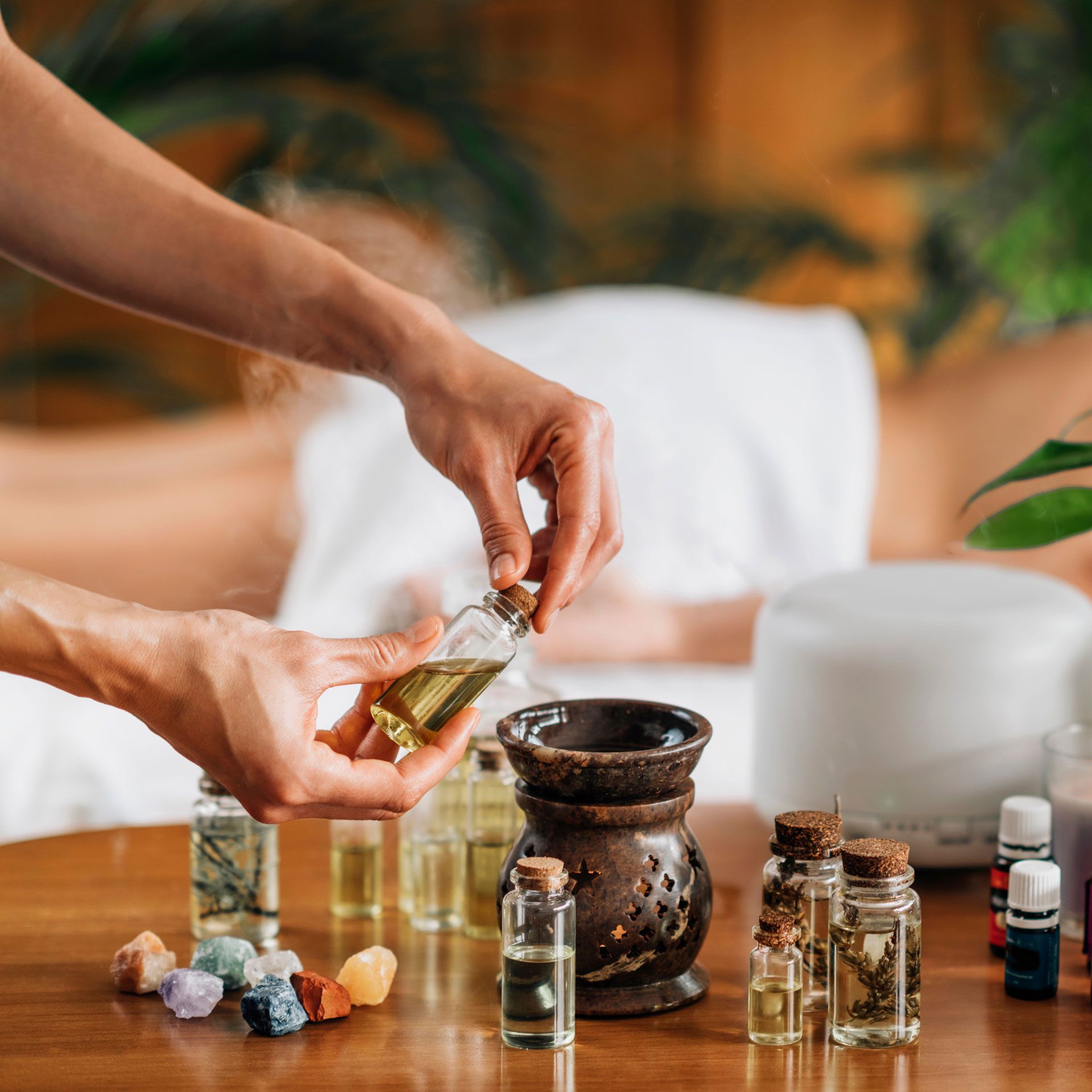 Person pouring oil into a diffuser, preparing for a massage. Various essential oil bottles on a table.