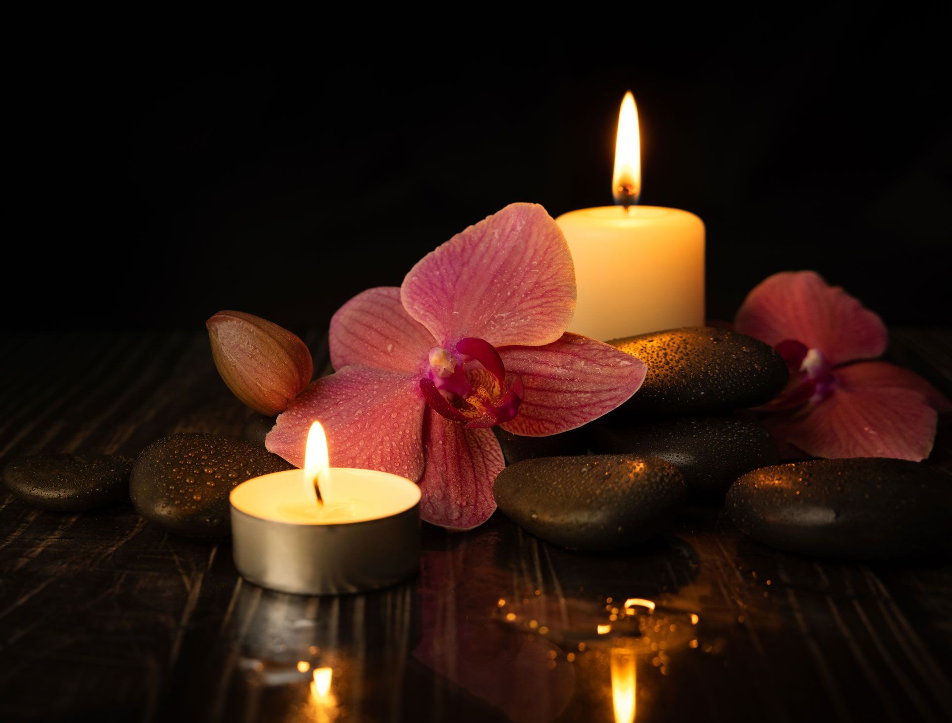 Two lit candles, pink orchid, and black stones on a dark, reflective surface.