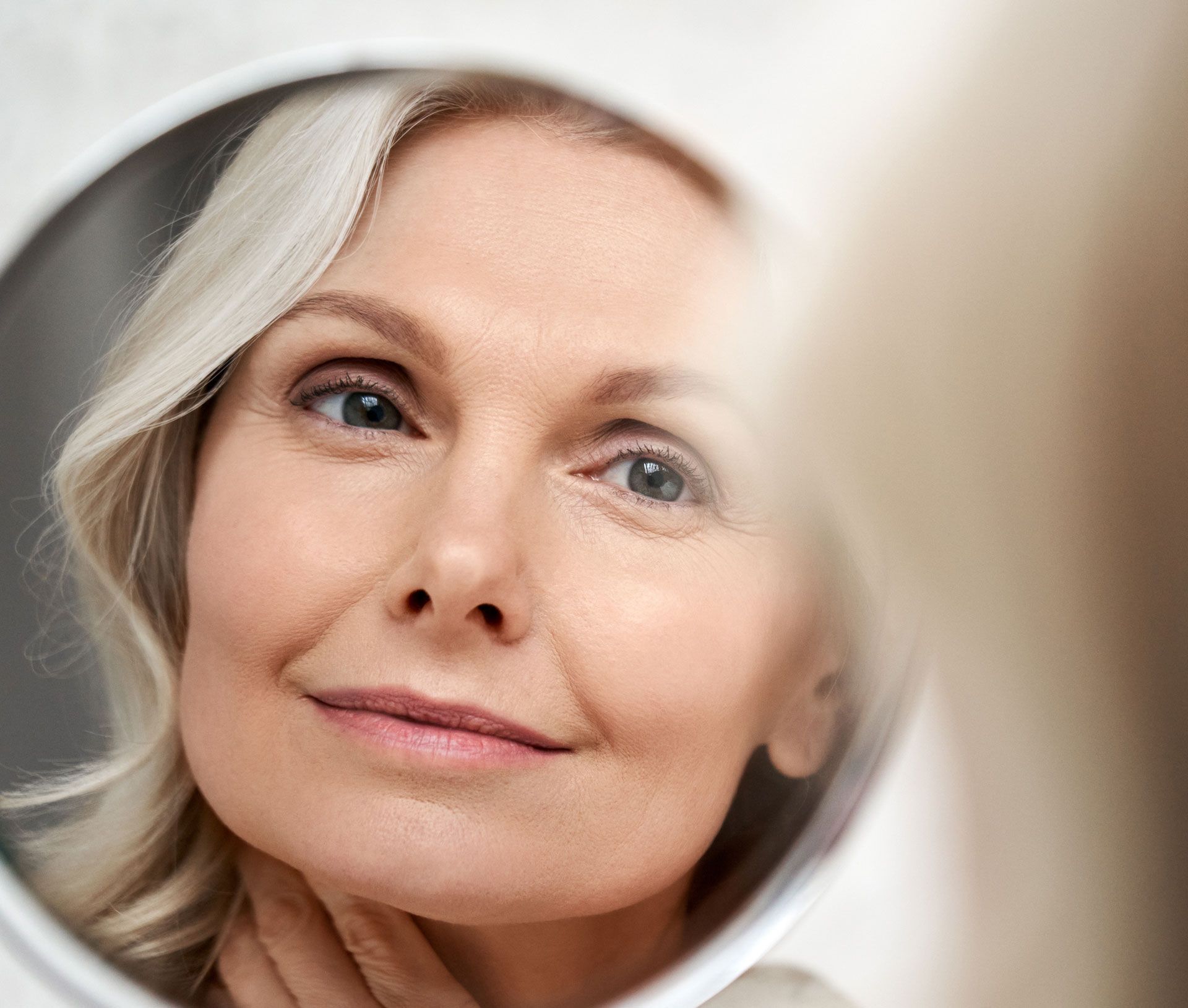Woman looking at her face in a circular mirror. She touches her neck. Light skin, blonde hair.