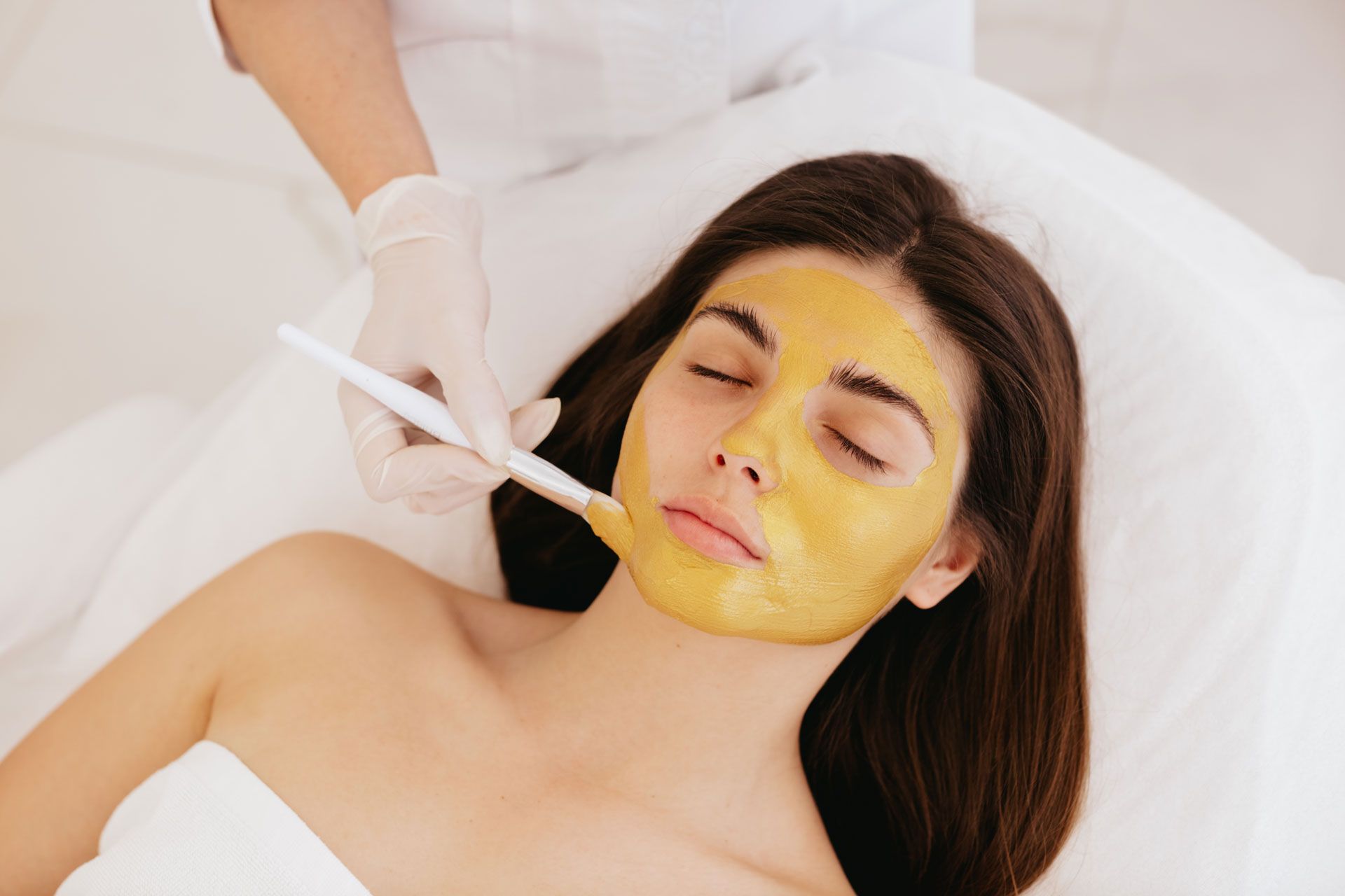 Woman receiving facial treatment with a golden mask applied by gloved hand.