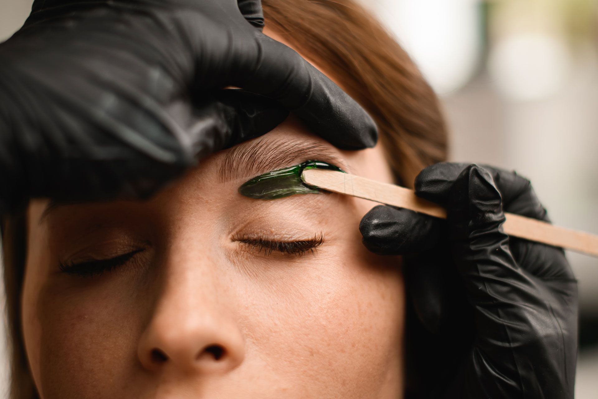 Eyebrow waxing: A person's eyebrow being waxed by gloved hands with a wooden applicator.