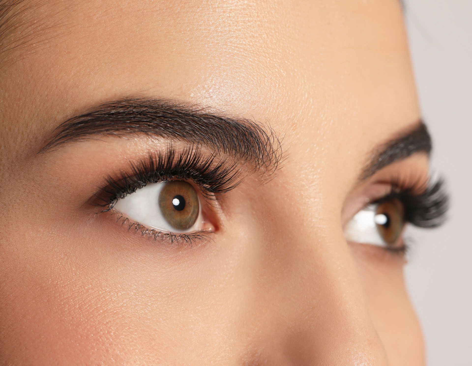 Close-up of brown eyes with thick, dark eyelashes and well-groomed, filled-in eyebrows. Skin has a warm, healthy tone.