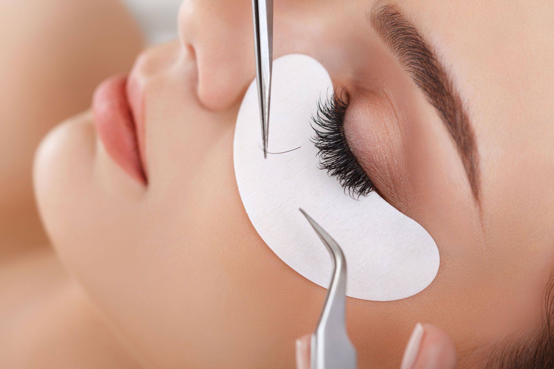 A person having eyelash extensions applied. They have an eye patch and tweezers being used on their lashes.