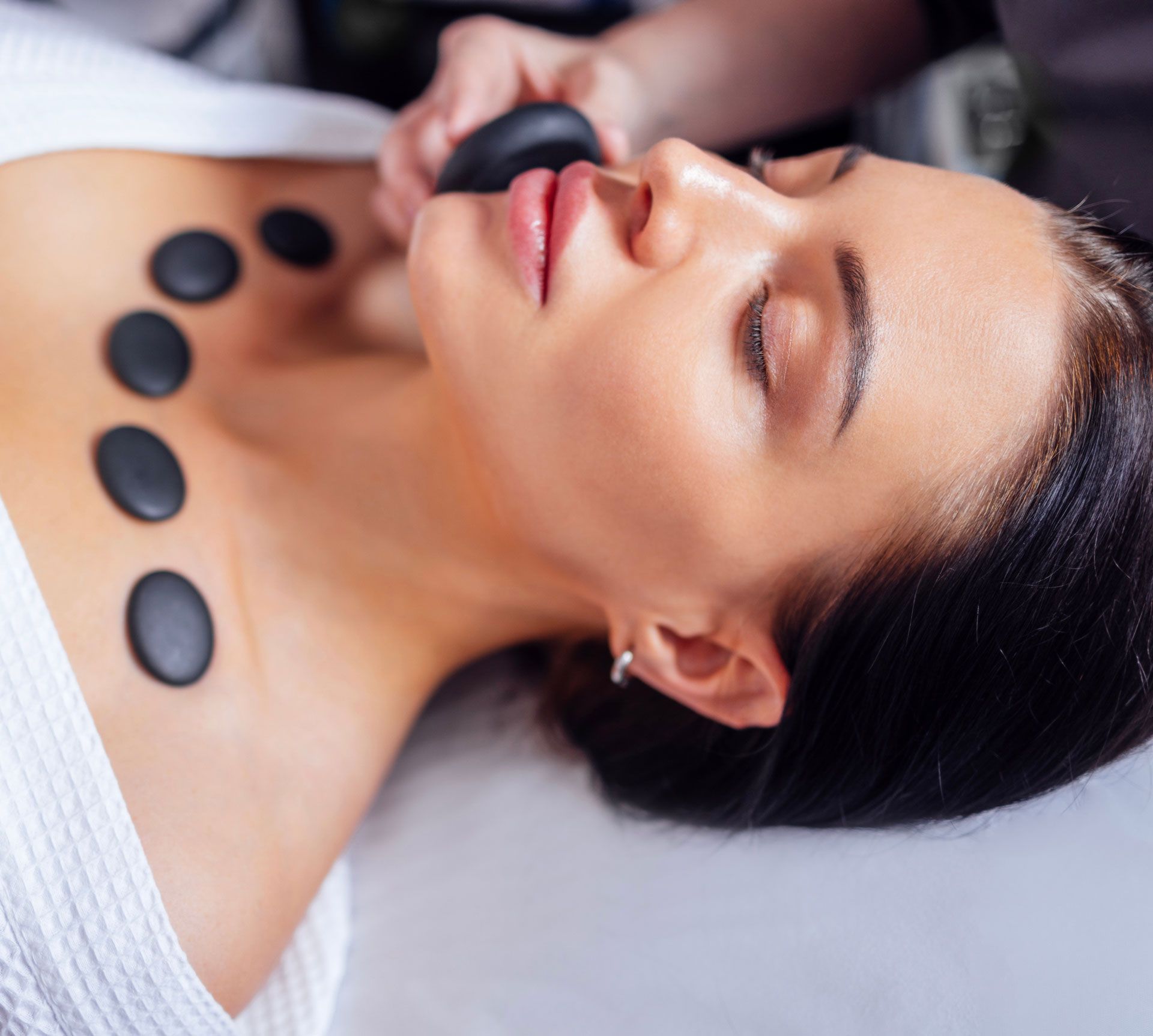 Woman receiving hot stone massage on her neck and face in a spa setting.