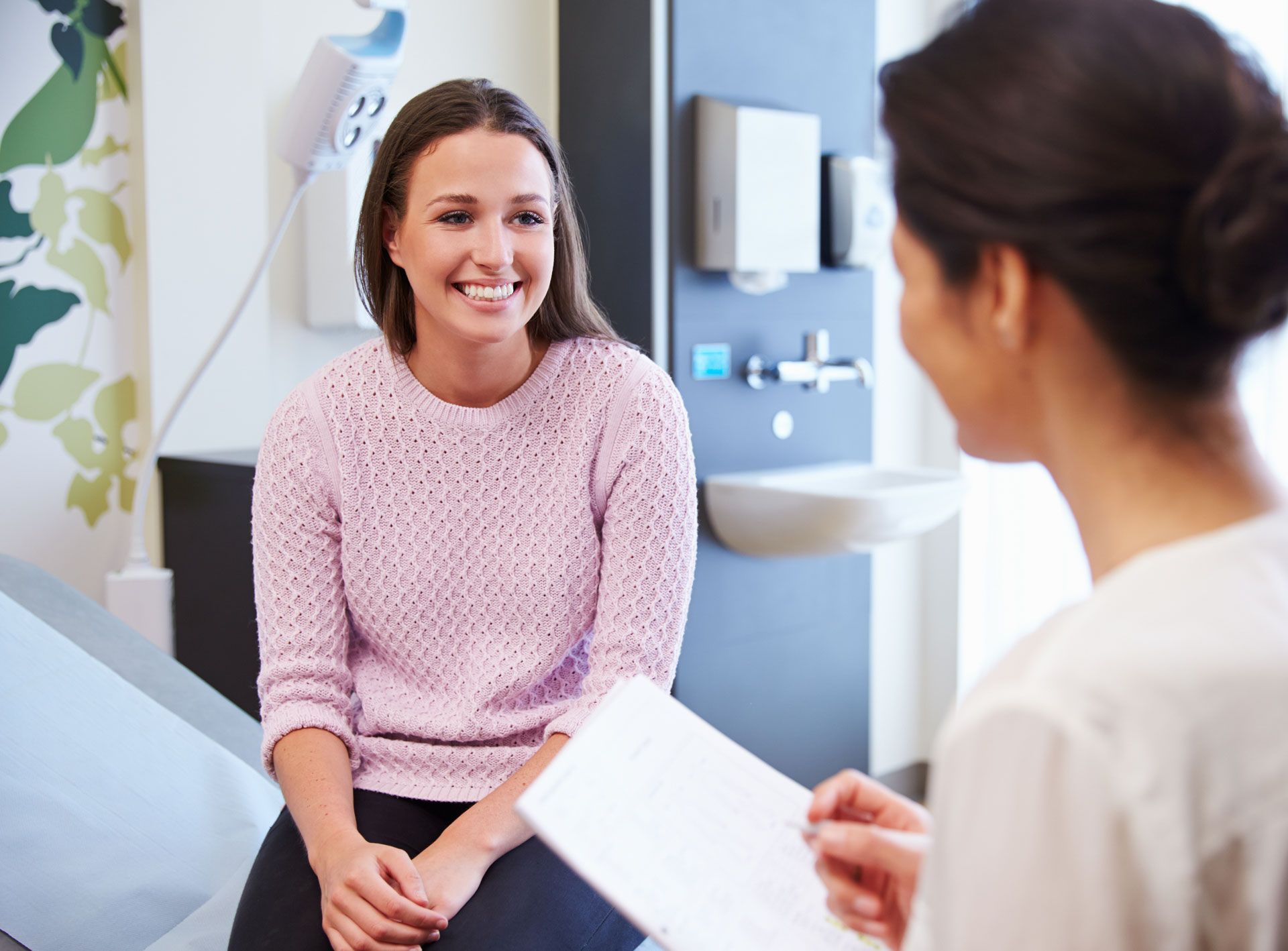 Woman smiling, seated, talking with a medical professional holding a paper in an exam room.