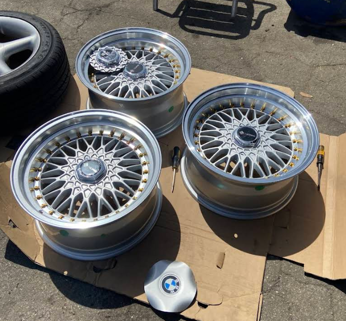 Three silver mesh-style wheels with BMW center caps arranged on cardboard on a paved surface.