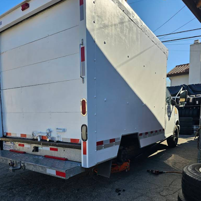 A white box truck parked outdoors with a rear tire removed and a floor jack positioned underneath the axle.