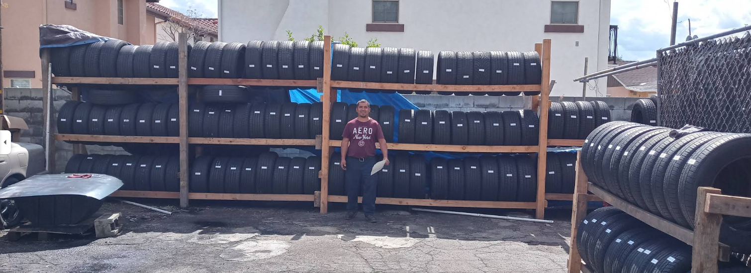 A person stands in front of a large, three-tiered wooden rack stocked with rows of stacked black tires outdoors.