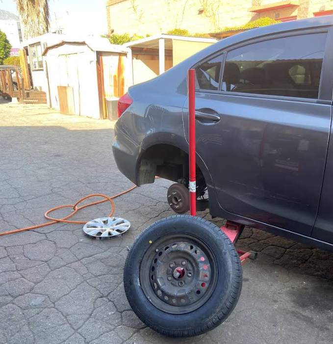 A gray car is raised on a red jack stand outdoors, with its rear tire removed and placed on the pavement nearby.