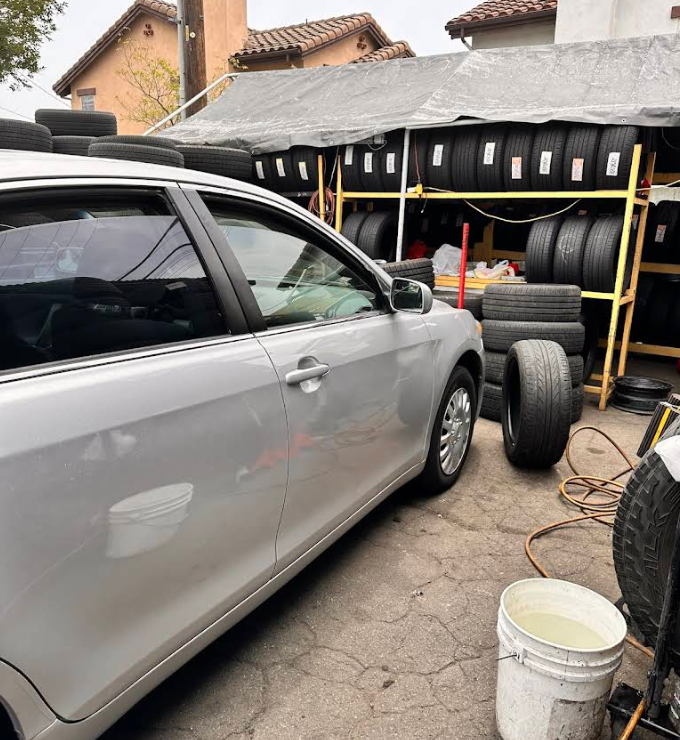 A silver sedan parked outdoors next to a makeshift tire rack stocked with various black tires.