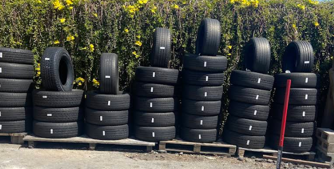 Several stacks of black rubber tires arranged on wooden pallets in front of a green hedge.