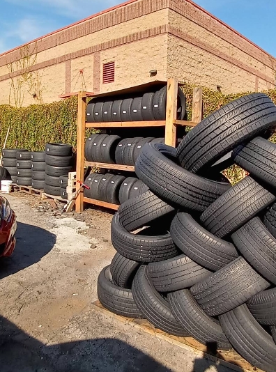 A large pile of black rubber tires stacked on a wooden rack and on the ground outside a brick building.