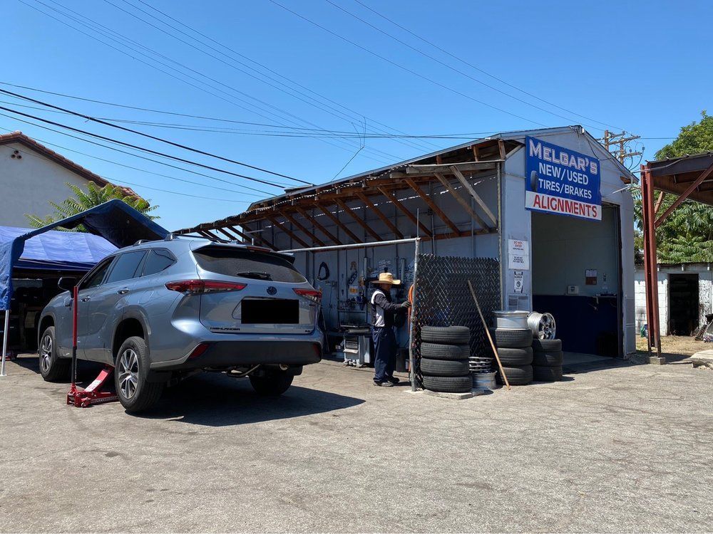 A silver SUV lifted by a red floor jack outside an auto repair shop with a worker standing near stacks of tires.