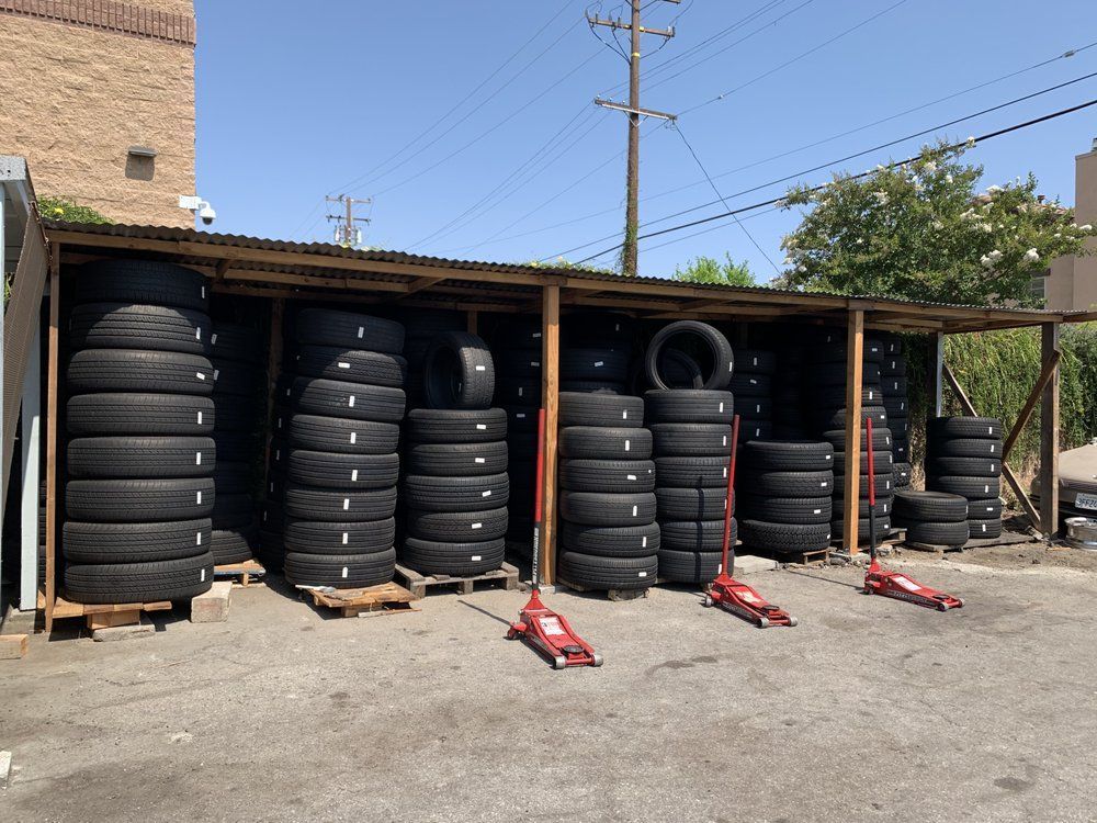 Rows of stacked tires are organized under a covered outdoor structure, with several red hydraulic jacks parked in front.