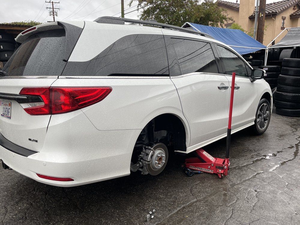 A white minivan parked on a concrete surface, raised by a red floor jack with its rear passenger-side wheel removed.