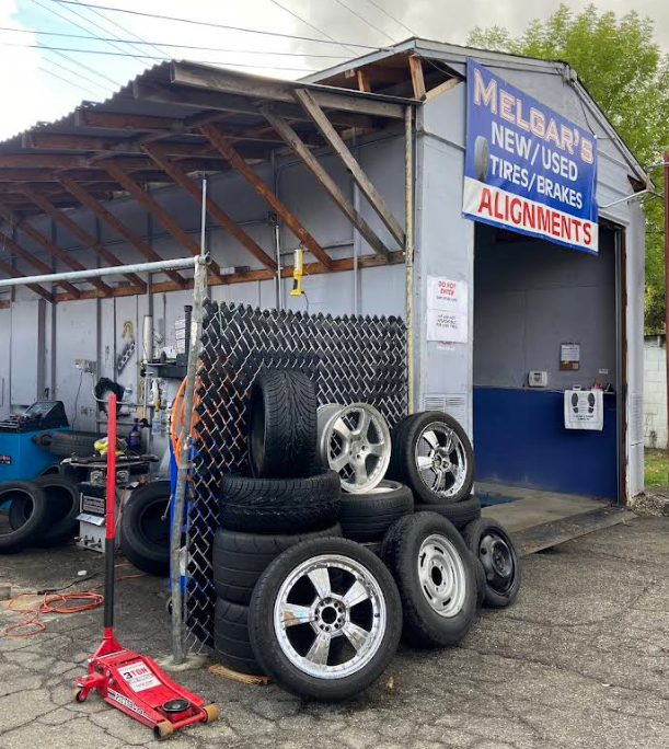 A tire shop exterior with a stack of tires and wheels, a red floor jack, and a sign advertising tires and alignments.