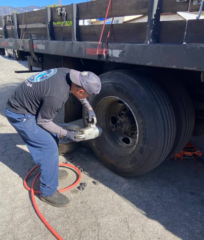 A mechanic uses a pneumatic impact wrench to remove lug nuts from the rear dual wheels of a large flatbed truck outdoors.