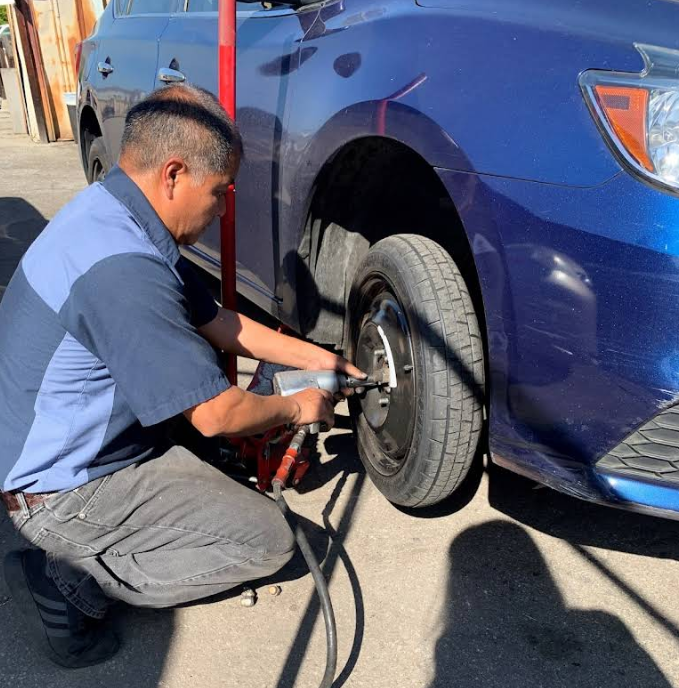 A technician uses a pneumatic impact wrench to remove lug nuts from the front wheel of a blue car.