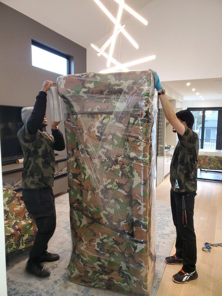 Two movers in camo shirts lift a large, wrapped piece of furniture in a modern home.