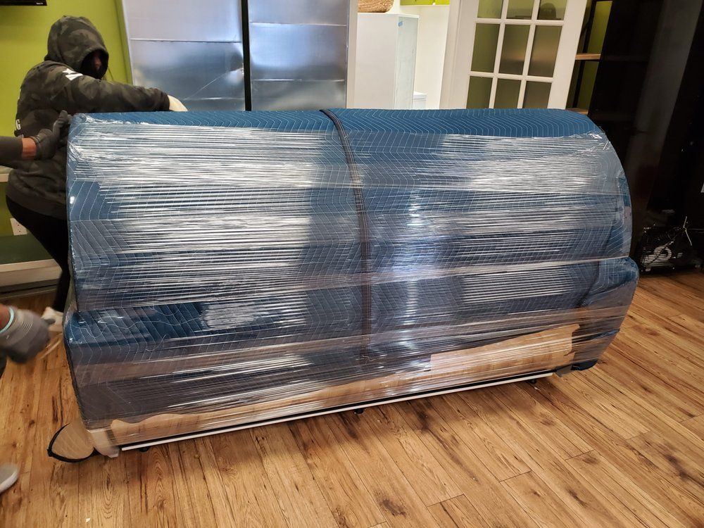 Two people moving a blue couch wrapped in plastic on a hardwood floor.