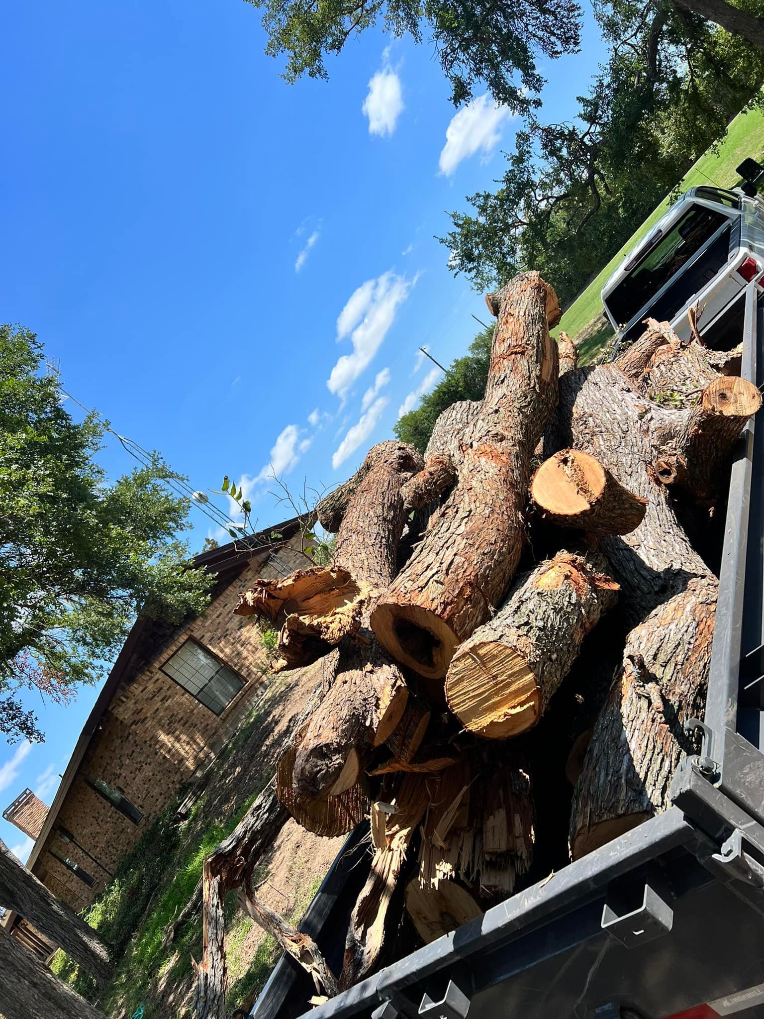 Logs piled in a truck bed, in front of a brick house under a blue sky.