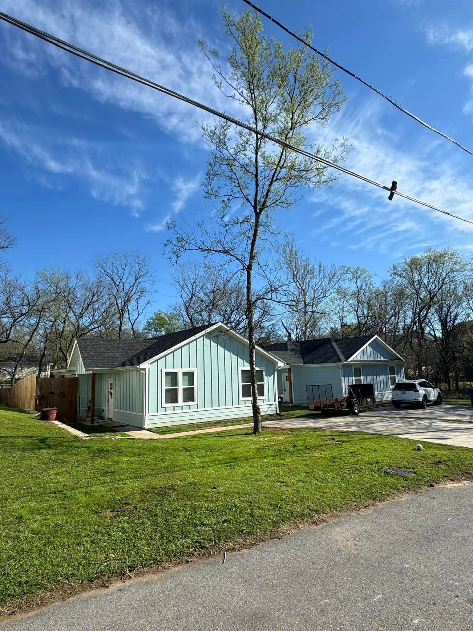 A light blue house with a dark roof and a tall tree in front on a sunny day.