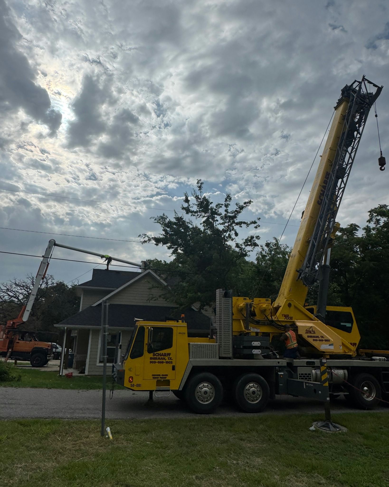 Yellow crane near a house with an elevated boom reaching over the roof, on a cloudy day.