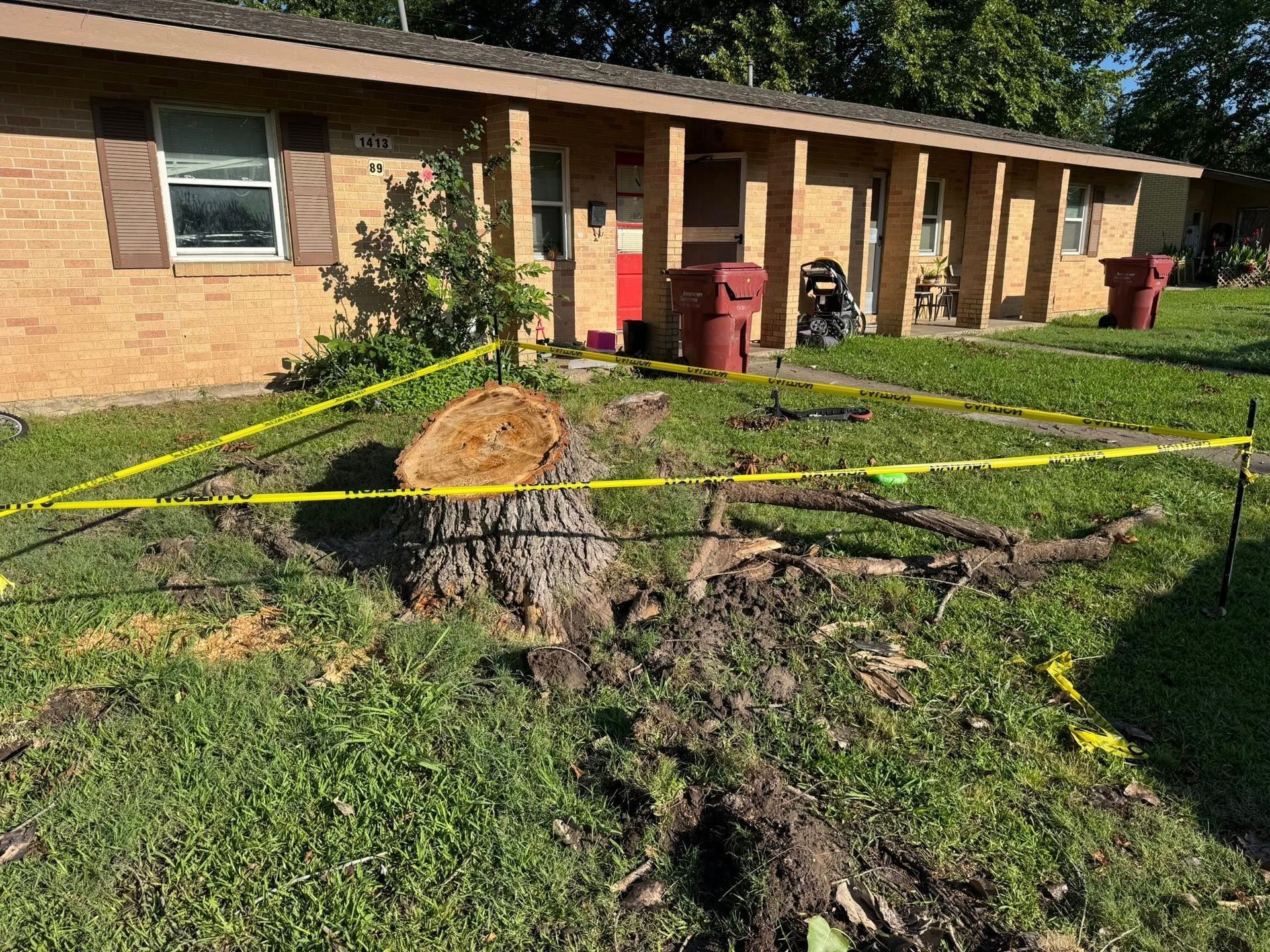 A tree stump with caution tape in front of a brick apartment building on a grassy lawn.