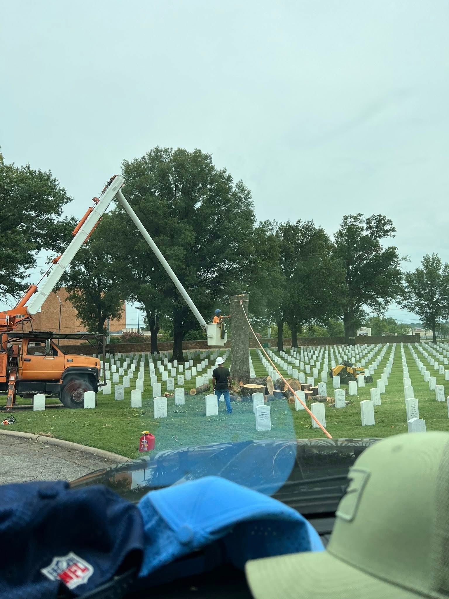 Tree trimming in a military cemetery with an aerial lift, rows of headstones, and workers.