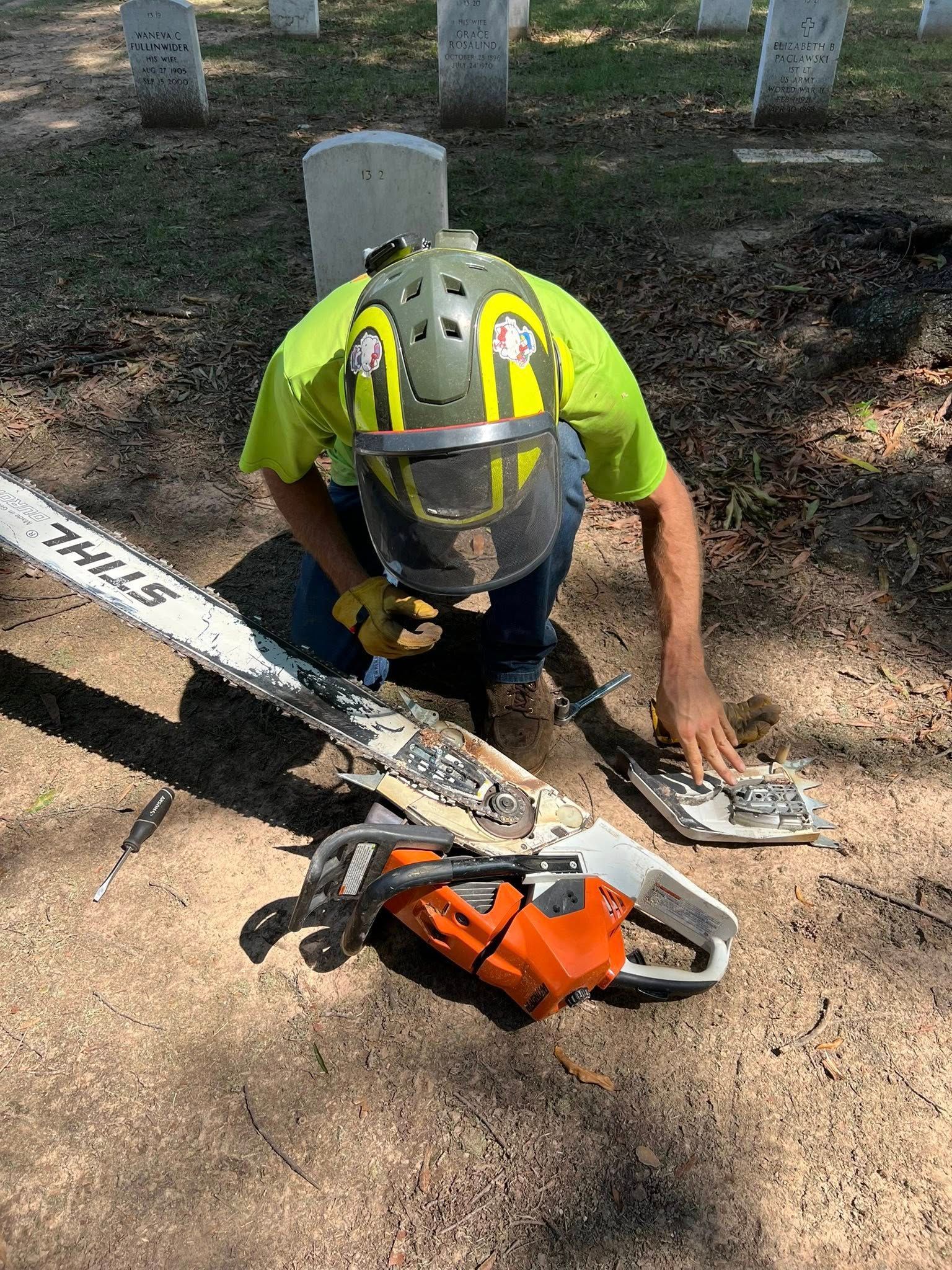 Person with chainsaw, wearing protective gear, cleaning the saw in a graveyard.