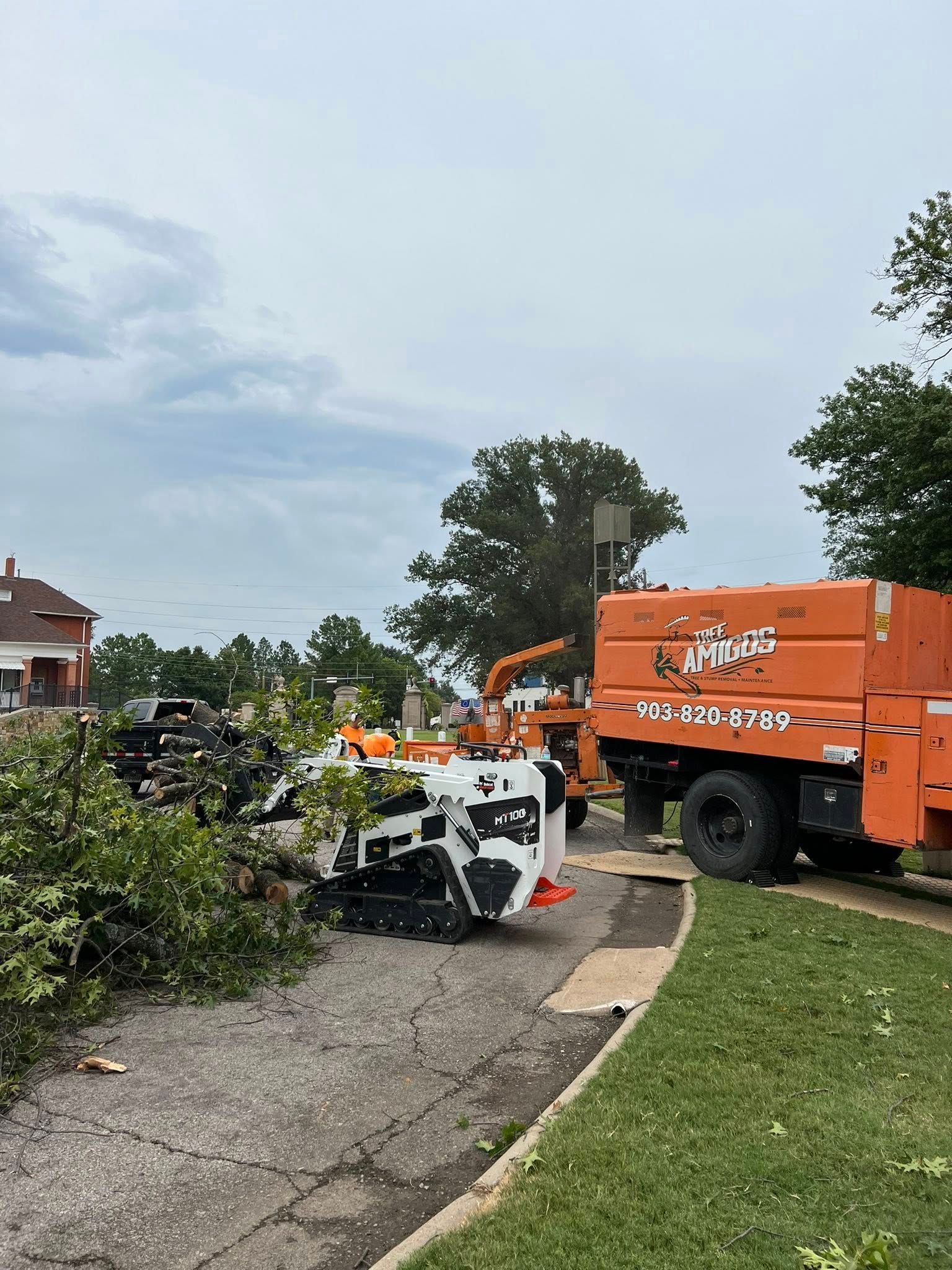 Orange wood chipper and truck, processing branches on a residential street with a cloudy sky overhead.