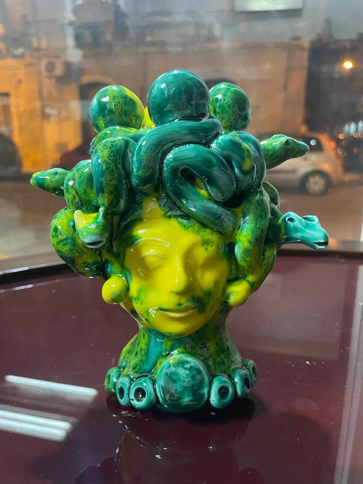 Scultura in ceramica raffigurante una testa di Medusa, gialla e verde, per interni con sfondo urbano.