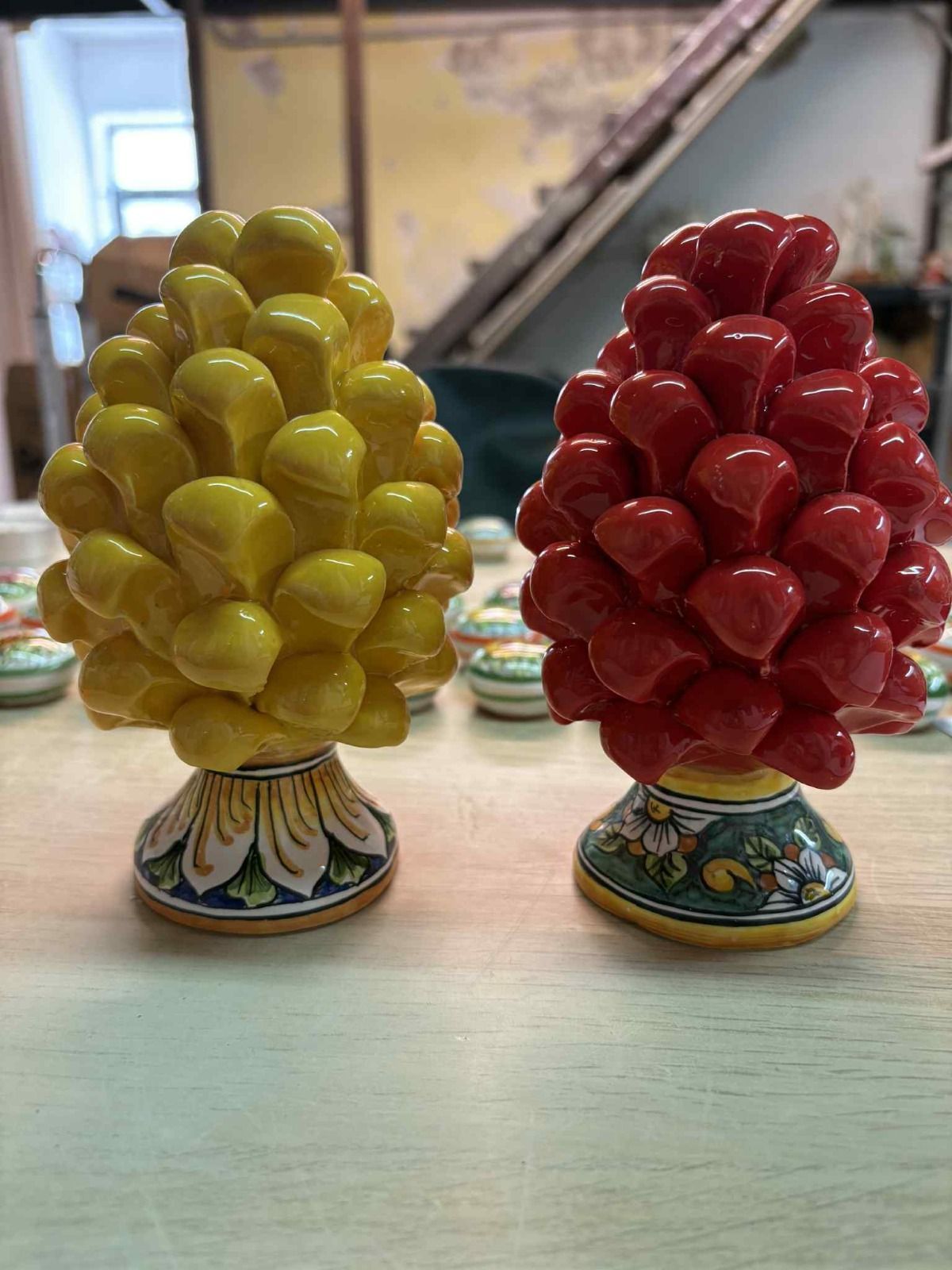 Pigne di ceramica gialle e rosse su un tavolo di legno, con basi a motivi floreali e uno sfondo di oggetti vari.
