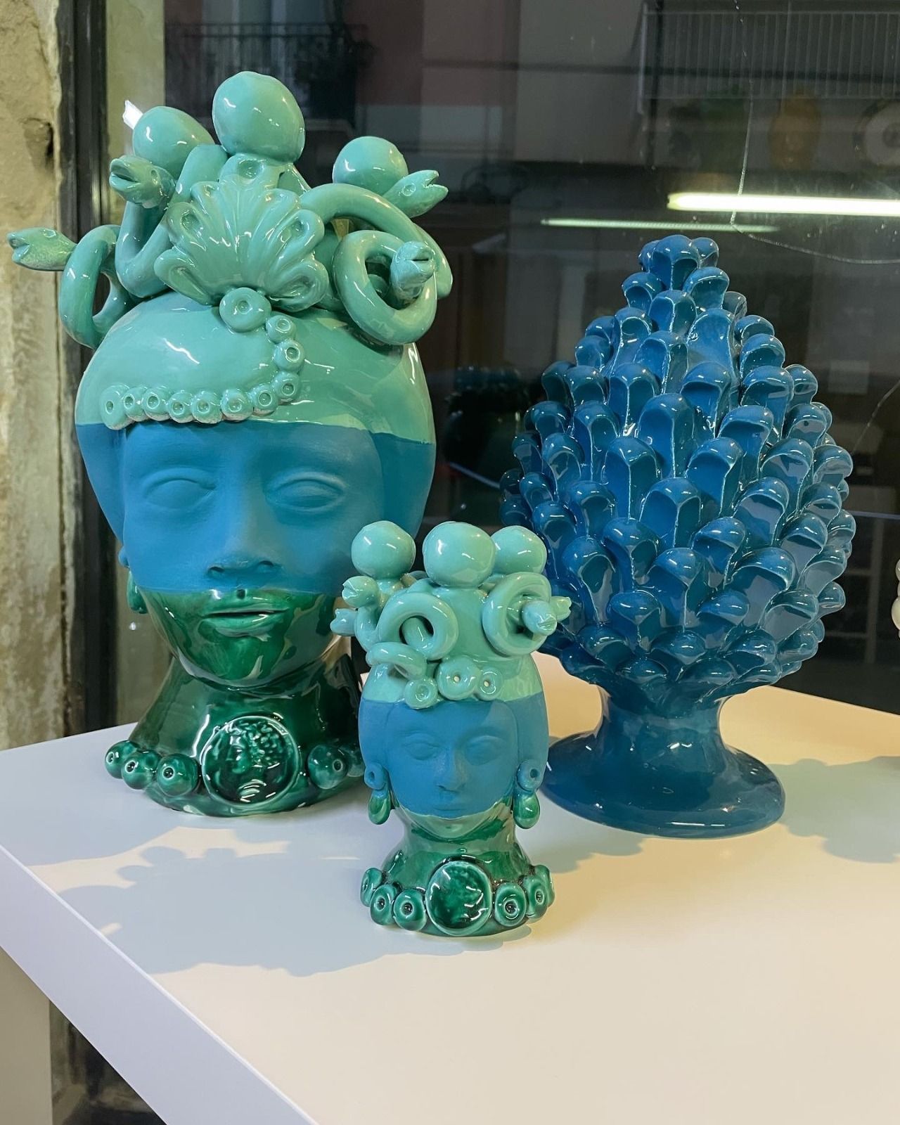 Tre sculture in ceramica color turchese e verde: due vasi a forma di viso e una pigna su una mensola bianca.