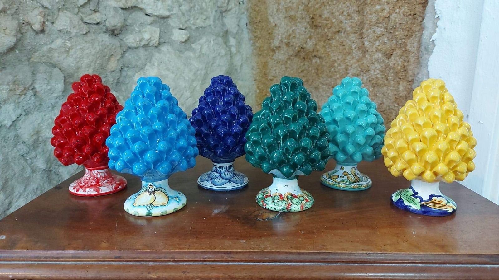 Sculture di pigne in ceramica colorata su una superficie di legno, appoggiate a un muro di pietra.
