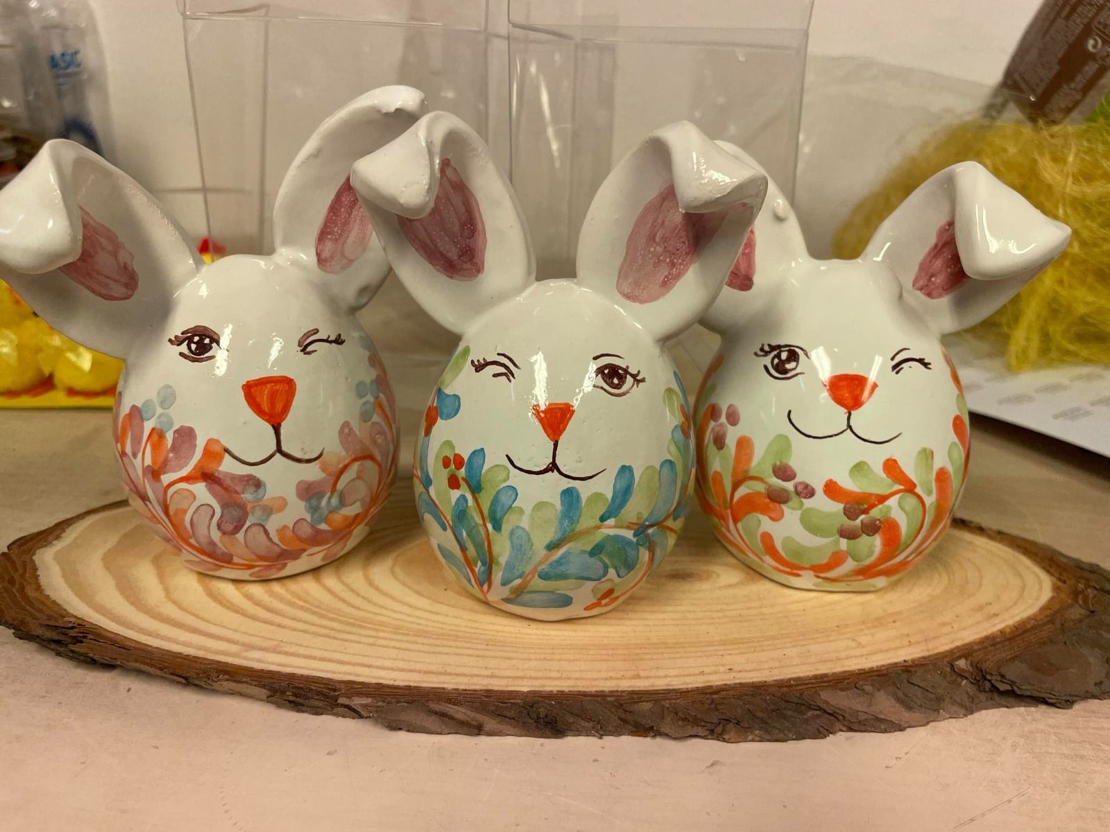 Tre uova di Pasqua in ceramica raffiguranti coniglietti con facce dipinte e motivi floreali su una lastra di legno.