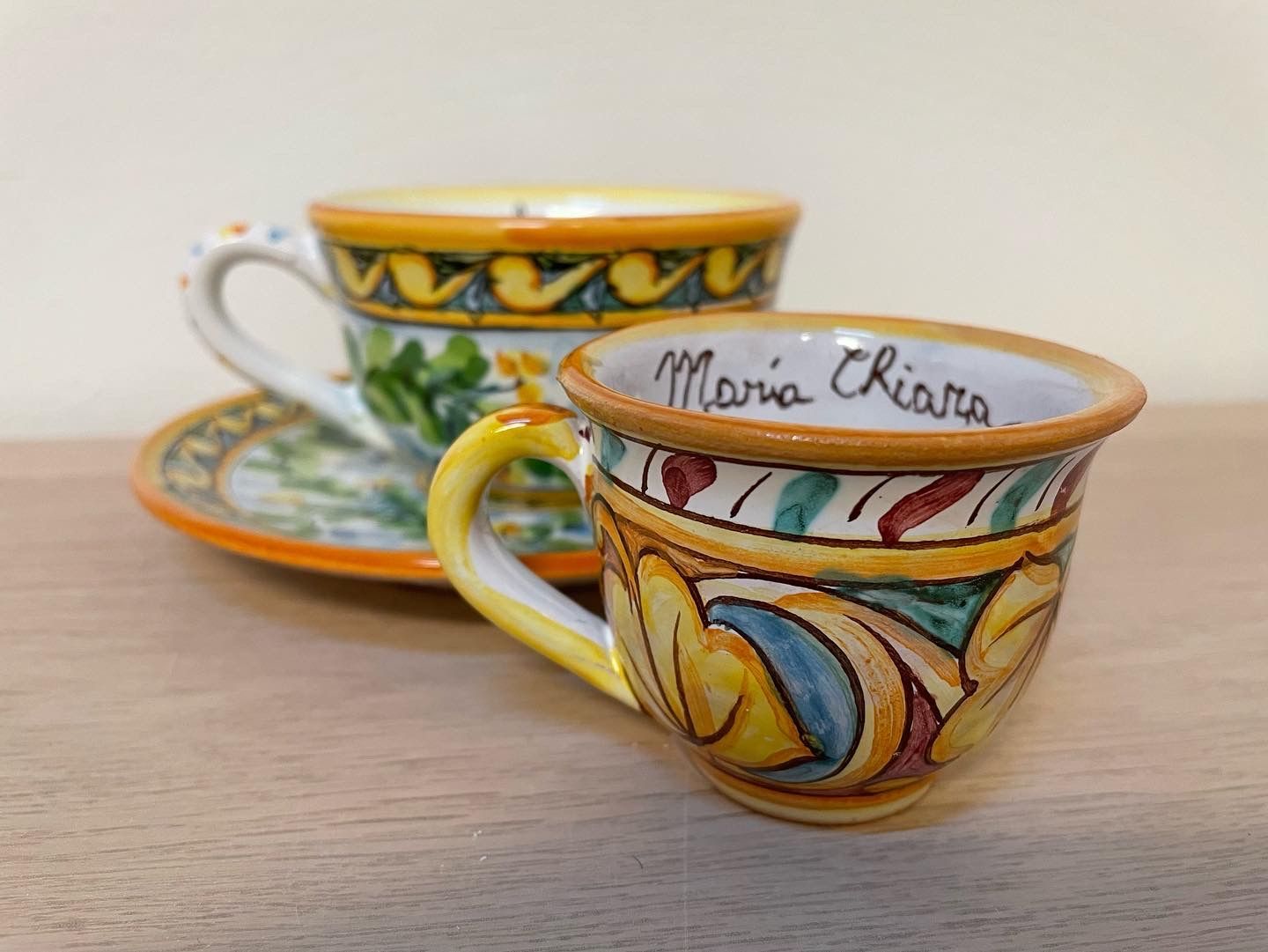 Due tazzine da caffè in ceramica dipinte a mano; una con piattino. Motivi floreali gialli, blu e arancioni. Scritta 