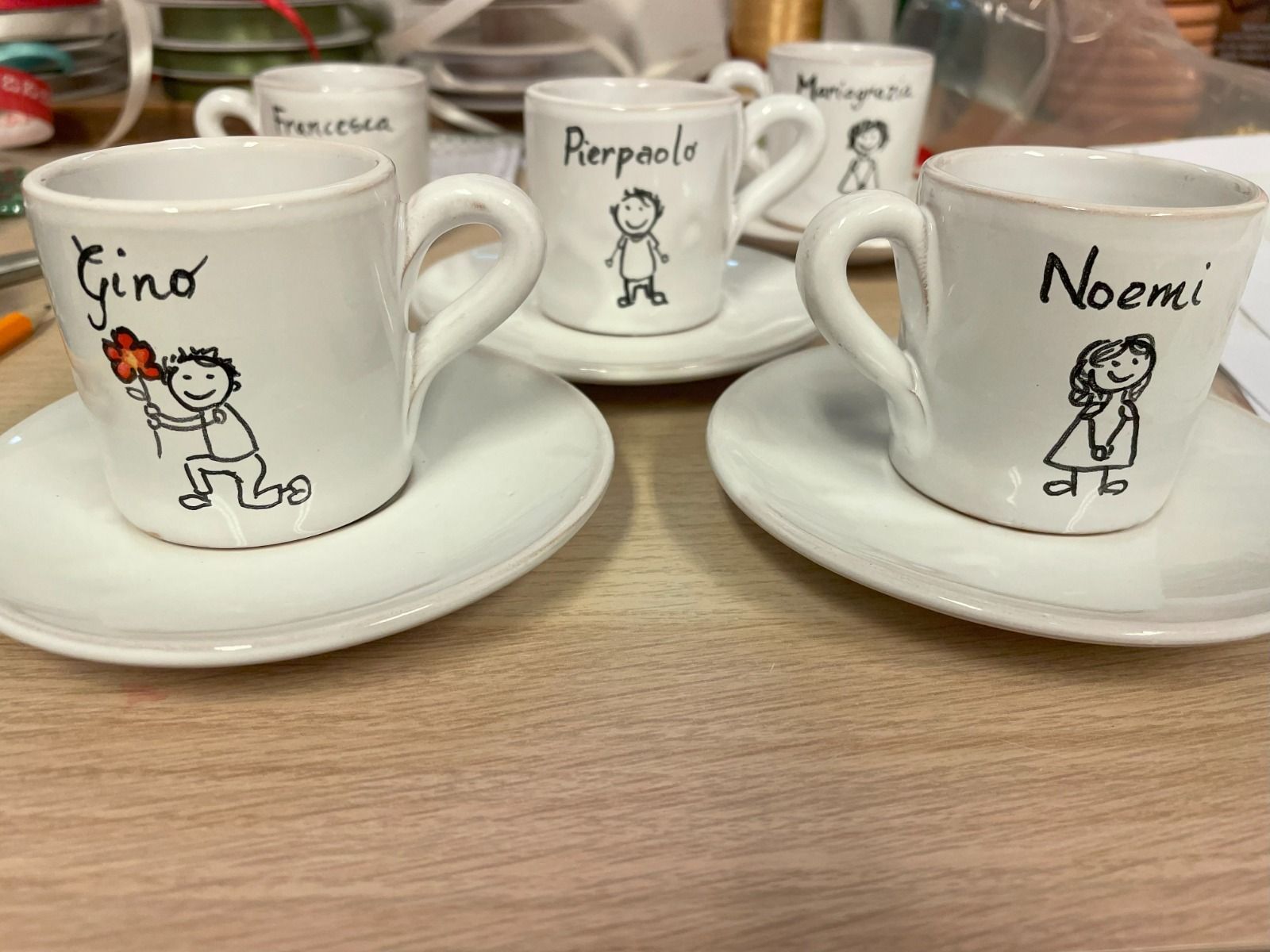 Cinque tazzine da caffè bianche con piattini, ciascuna disegnata a mano con il nome di una persona e un'immagine di un cartone animato.