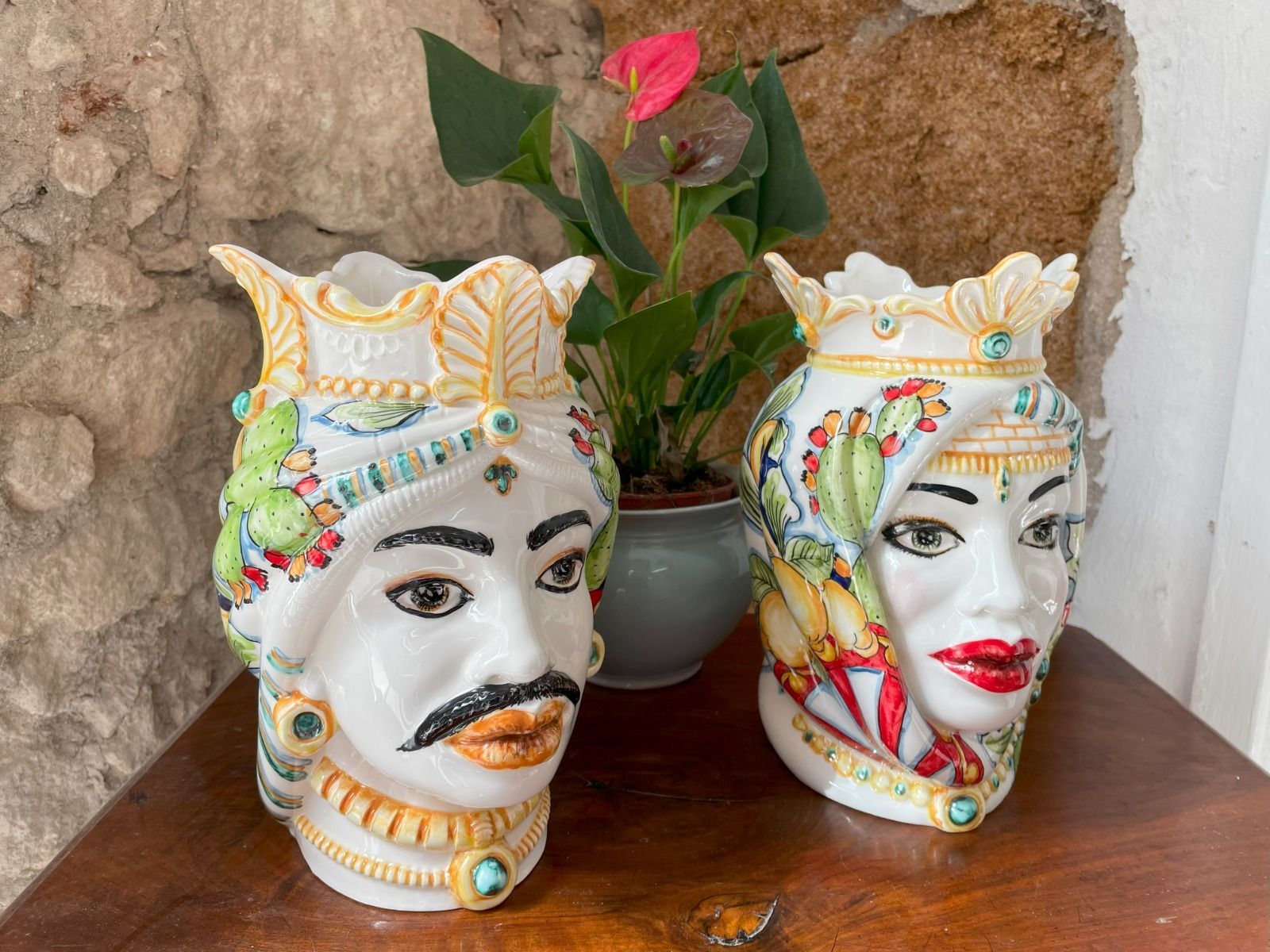 Coppia di vasi in ceramica ornati, raffiguranti una testa maschile e una femminile, con dettagli colorati, di fronte a un muro di pietra.